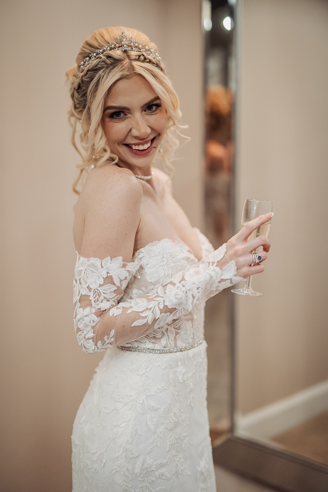 A smiling bride in an off-the-shoulder lace wedding dress holds a glass of champagne at Ashes Barn in Staffordshire. She has blonde hair styled with a jeweled headpiece and stands in front of a mirror, glowing on her wedding day.