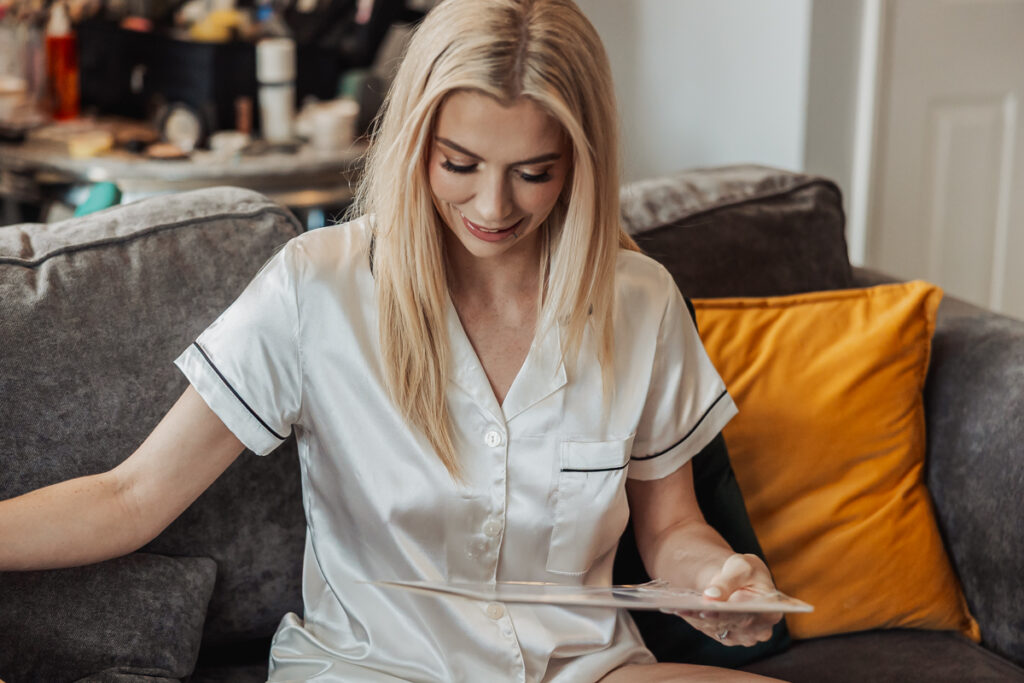 A woman with long blonde hair wearing white satin pajamas sits on a gray couch with yellow cushions, smiling and looking down at a wedding photo in her hand. A table with various items is in the background at an Ashes Barn in Staffordshire.