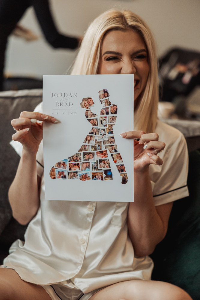 A woman in pajamas smiles and holds up a wedding poster featuring a silhouette made of small couple photos and the words “Jordan & Brad.” She is sitting on a couch at Ashes Barn, with another person’s blurred legs in the background.