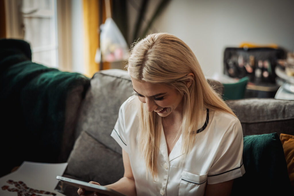 A woman with long blonde hair, wearing a white satin shirt, sits on a couch in a cozy Staffordshire living room, looking down at a tablet and smiling—perhaps browsing wedding ideas for Ashes Barn.