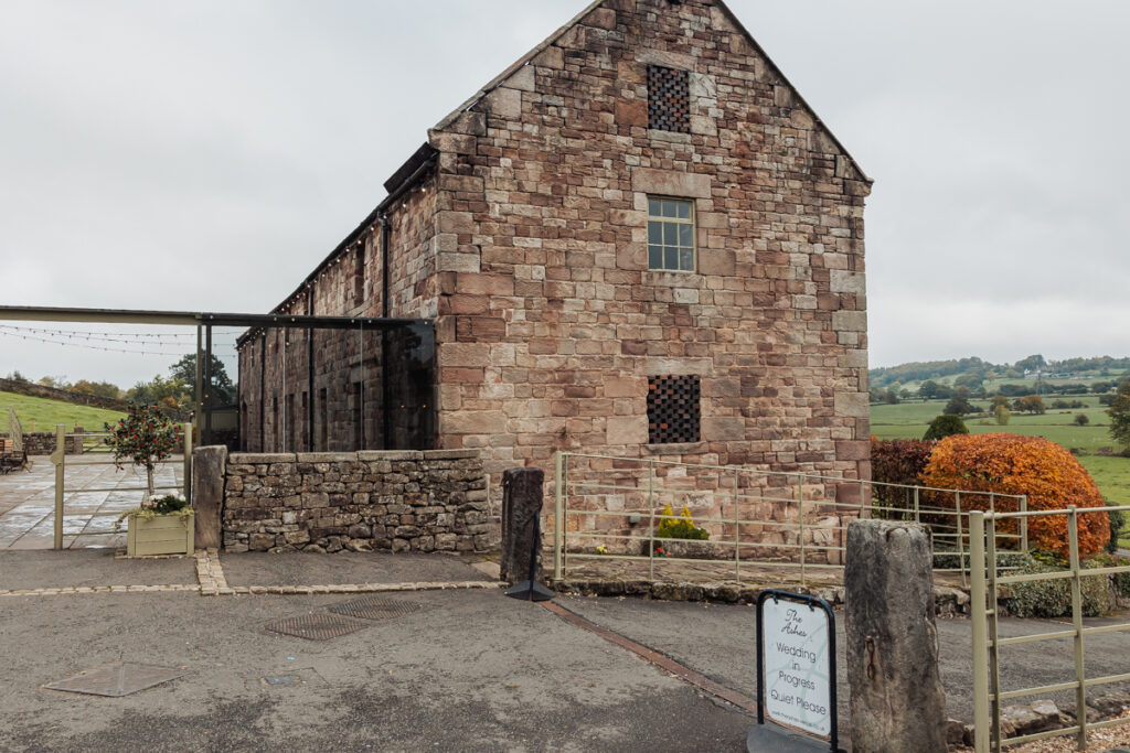 Ashes Barn, a large old stone building with small windows, sits by a paved area in Staffordshire’s countryside. Green fields and a distant hill lie beneath a cloudy sky—a charming wedding venue, marked by a sign and metal railing in the foreground.