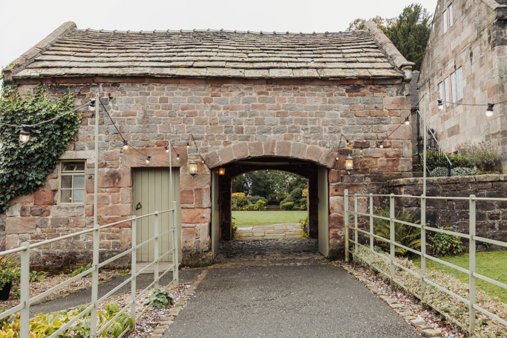 Stone building with a tiled roof and arched passageway at Ashes Barn, Staffordshire, lanterns on either side, string lights overhead, a pale green door to the left, and a view through to a garden—perfect for a memorable wedding backdrop.