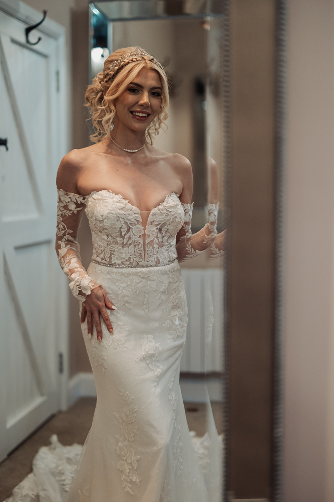 A smiling bride in an elegant, off-the-shoulder lace wedding dress and pearl necklace stands indoors at Ashes Barn in Staffordshire, looking at her reflection in a mirror on her wedding day.