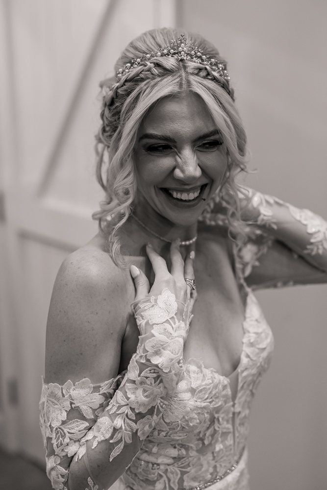 A joyful bride in a lace wedding dress and jeweled headpiece smiles brightly, holding her hands to her chest and neck. Captured in black and white at Ashes Barn, Staffordshire, the photo radiates her wedding happiness and excitement.