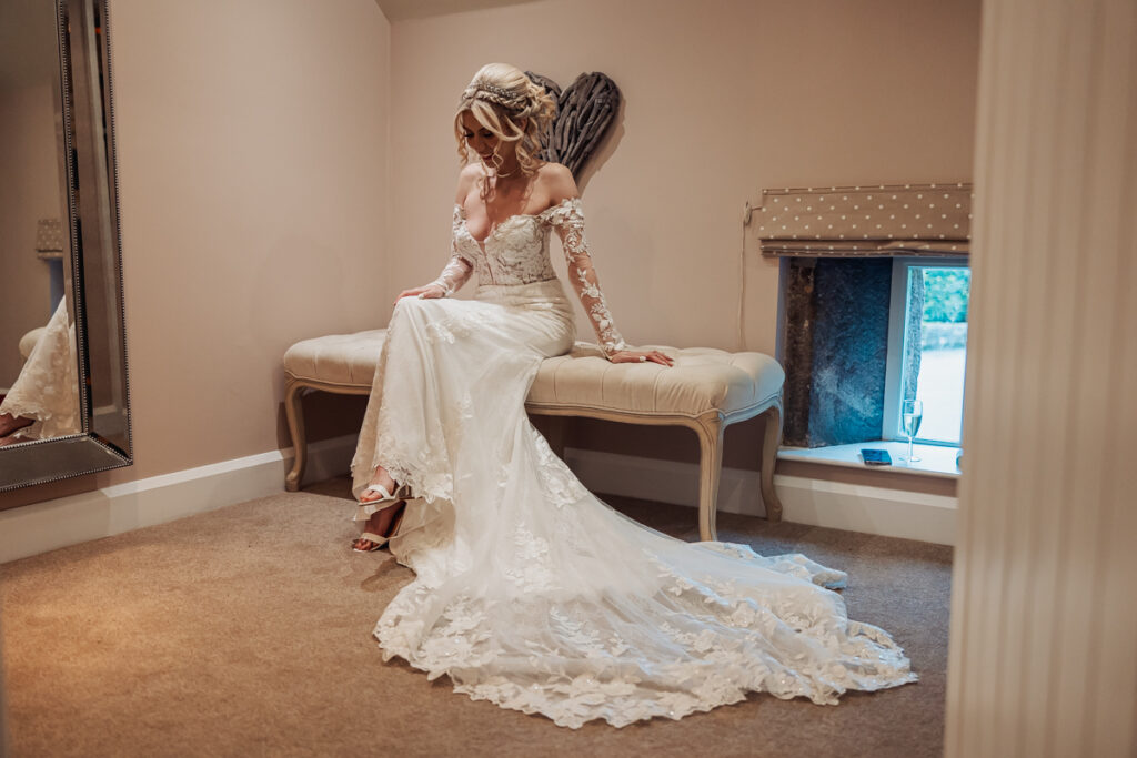 A bride in a long, lace wedding dress sits on a bench in a softly lit room at Ashes Barn in Staffordshire, looking down as she adjusts her sandal. The train of her dress flows gracefully over the carpeted floor.