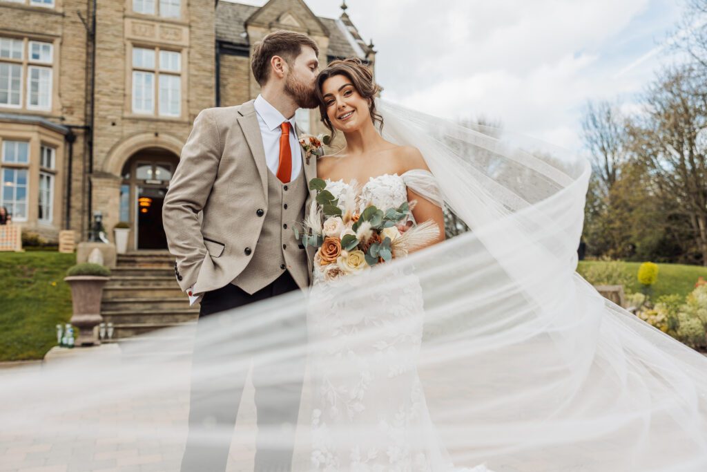 The bride, adorned in a flowing white gown with a long veil and holding a vibrant floral bouquet, stands beside the groom in a tan suit and red tie, as they share a tender kiss