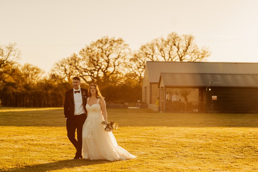 A couple in wedding attire stands together on a grassy field, bathed in warm, golden sunlight, with a barn in the background