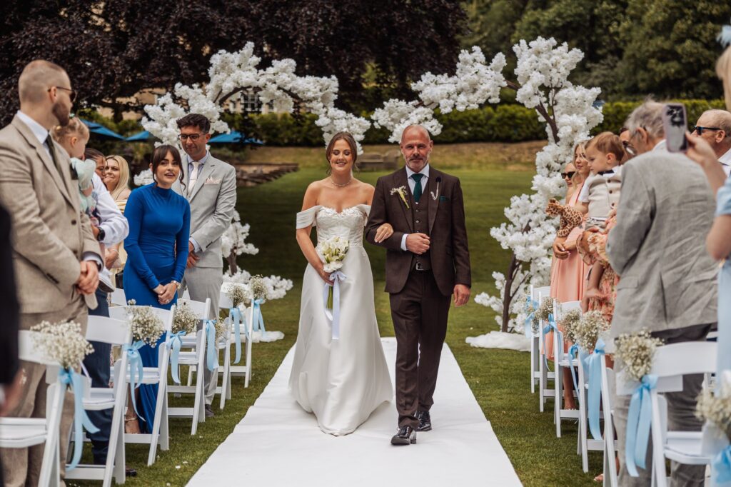 The bride walks the white aisle with her father, between blossom trees, at Wentbridge House Hotel.