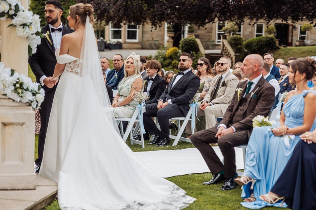 The bride at the front of the ceremony, with the stone frontage of Wentbridge House Hotel behind the seated guests.
