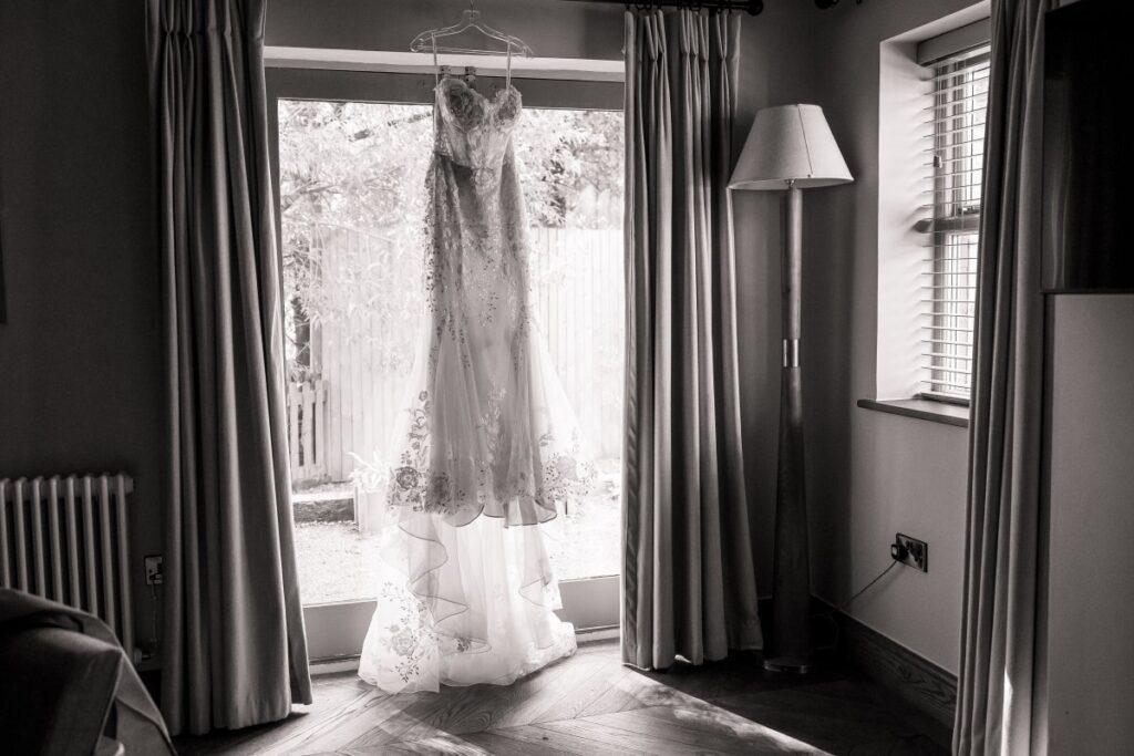 A lace wedding dress hangs in front of a glass door framed by curtains, with natural light streaming in—evoking the romantic charm of Bolton Abbey. A floor lamp and window with blinds are on the right in the softly lit room, reminiscent of the serene ambiance found at Tithe Barn.