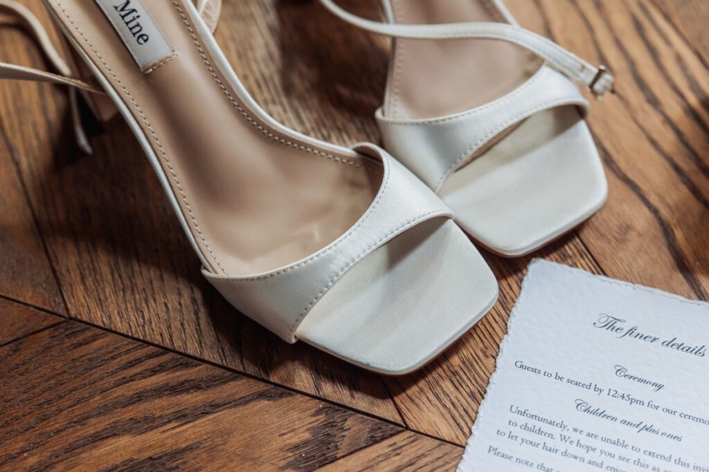 A pair of elegant white high-heeled sandals sits on a wooden floor next to a piece of paper with printed wedding details partially visible, hinting at a romantic celebration at the Tithe Barn, Bolton Abbey.