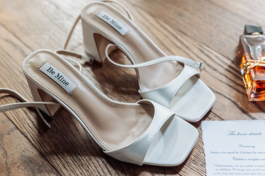 A pair of elegant white heeled sandals labeled Be Mine on the insole, placed on a wooden floor next to a perfume bottle and a partially visible printed invitation for a celebration at the Tithe Barn, Bolton Abbey.