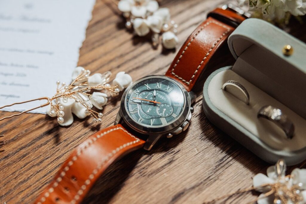A wristwatch with a brown leather band lies on a wooden surface next to a small box containing two rings and decorative pearl accessories, evoking the timeless elegance of Bolton Abbey’s historic tithe barn.