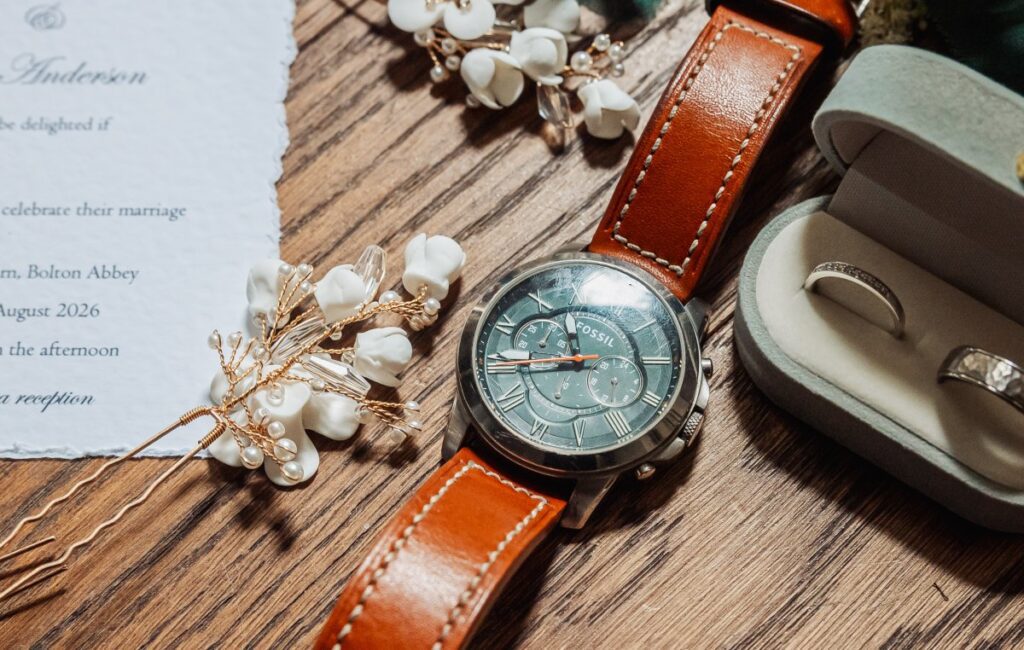 A brown leather wristwatch, a pair of rings in a box, and decorative white flowers rest on a wooden surface next to part of a wedding invitation, evoking the rustic elegance of Bolton Abbey’s Tithe Barn.