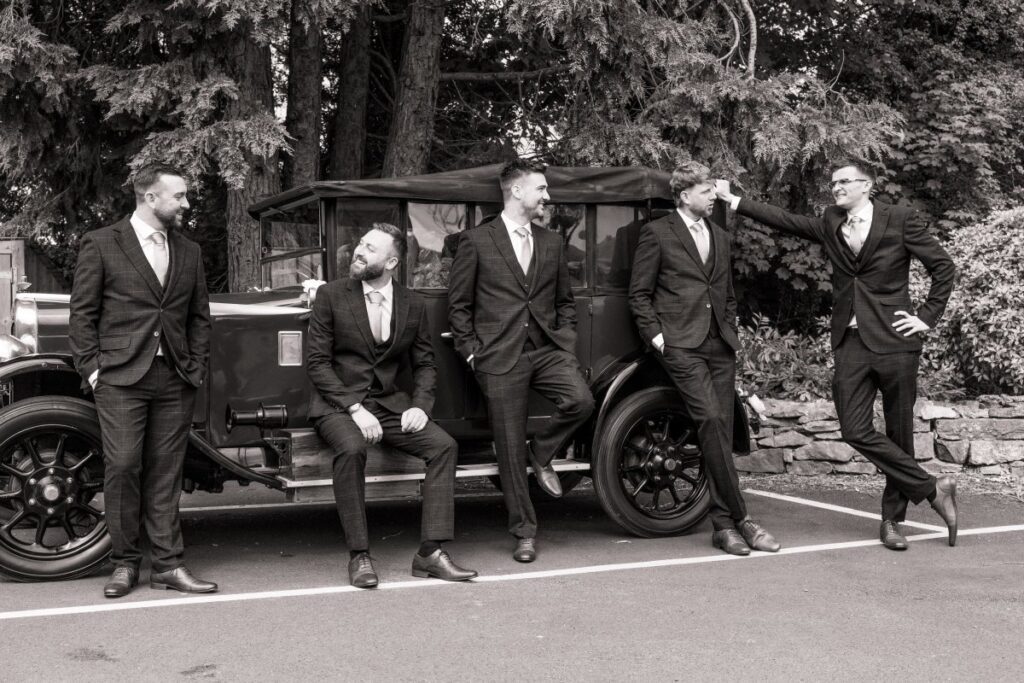 Five men in suits pose and laugh beside a vintage car in an outdoor setting, with trees and a stone wall near the Tithe Barn at Bolton Abbey in the background. The image is black and white.