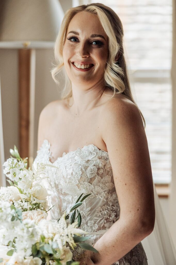 A smiling bride in a strapless lace wedding gown holds a bouquet of white flowers, standing indoors near a window with light streaming in at the Tithe Barn, Bolton Abbey.