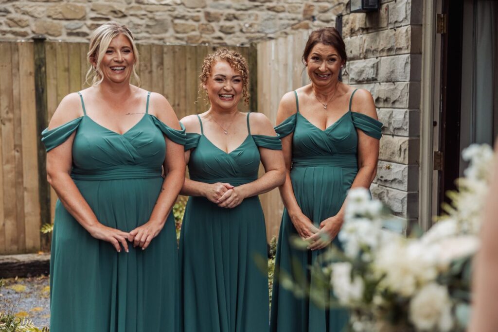 Three women in matching teal dresses smile and stand outside near a stone wall and wooden fence at Bolton Abbey, their hands clasped. A blurry bouquet of flowers is visible in the foreground, capturing a joyful moment beside the historic tithe barn.