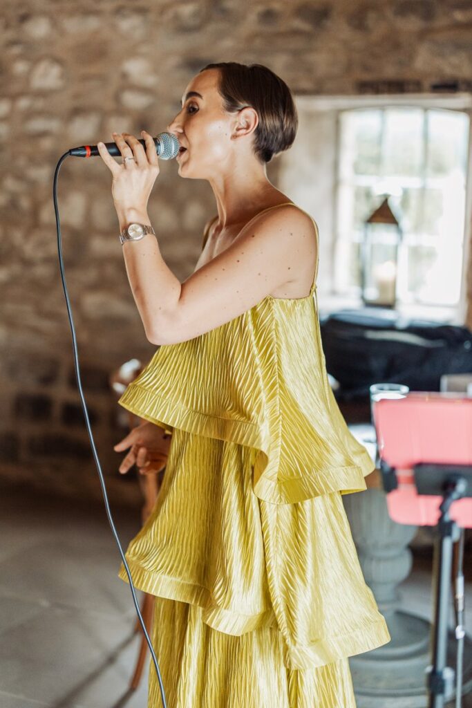 A person wearing a bright, pleated yellow dress sings into a microphone indoors at the Tithe Barn near Bolton Abbey, with natural light streaming through a window and musical equipment in the background.