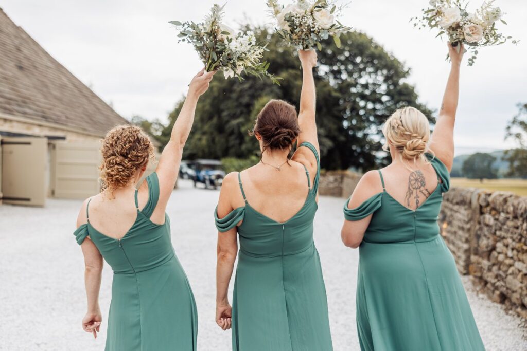 Three women in matching green dresses stand outdoors with their backs to the camera, each raising a bouquet of flowers in the air. They are on a gravel path by a stone wall near Bolton Abbey, with trees and a tithe barn visible in the background.