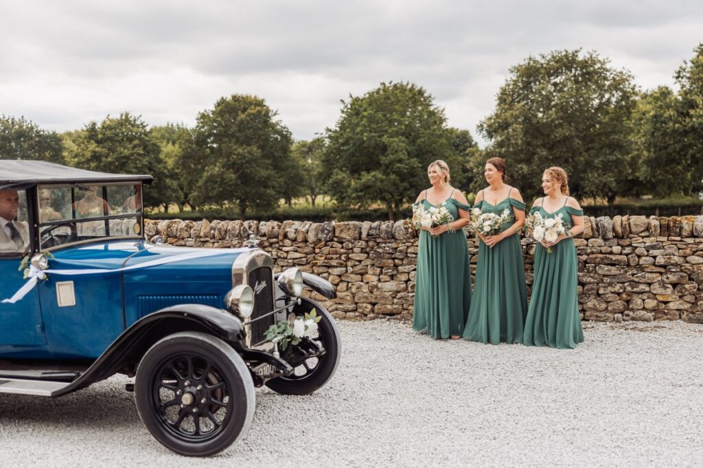 Three bridesmaids in green dresses stand by a stone wall holding bouquets, watching a vintage blue car decorated with white flowers arriving outside the Tithe Barn at Bolton Abbey on a cloudy day.