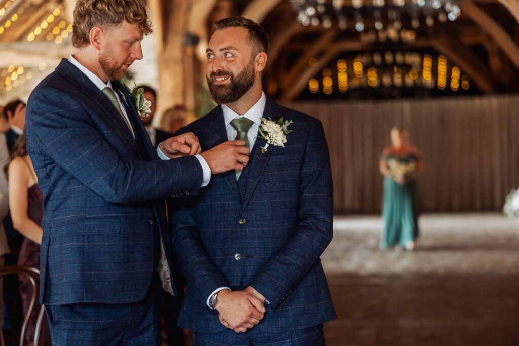In a warmly lit wooden hall, the groomsmen assist the groom with his boutonniere as guests gather in the background, creating a festive atmosphere at Tithe Barn, Skipton