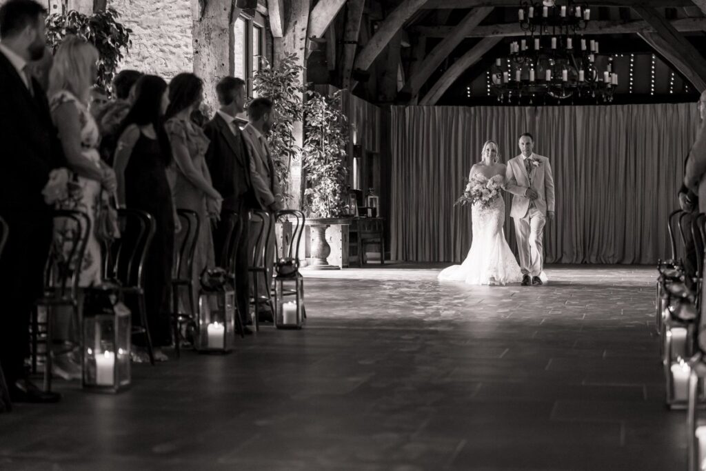 The bride walks down the aisle at Tithe Barn Bolton Abbey in black and white, guests standing as she approaches.