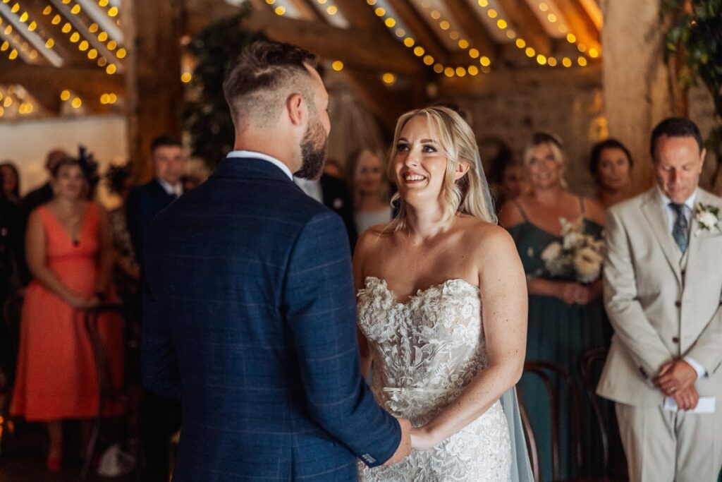 The bride smiles at her groom during the ceremony at Tithe Barn Bolton Abbey, beneath the fairy-lit beams.