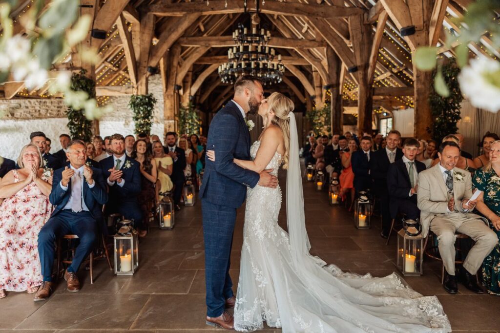 The first kiss as husband and wife at Tithe Barn Bolton Abbey, guests applauding from both sides of the aisle.