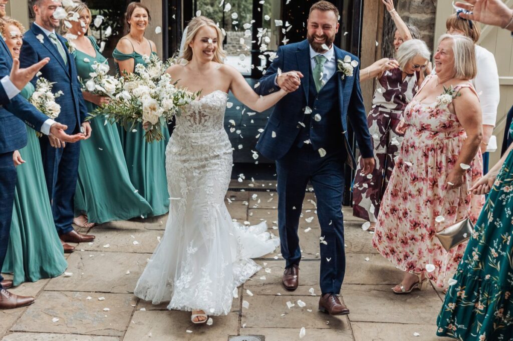 Confetti rains down as the bride and groom walk hand-in-hand through a stone courtyard, surrounded by smiling guests in vibrant dresses and suits at Tithe Barn, Skipton
