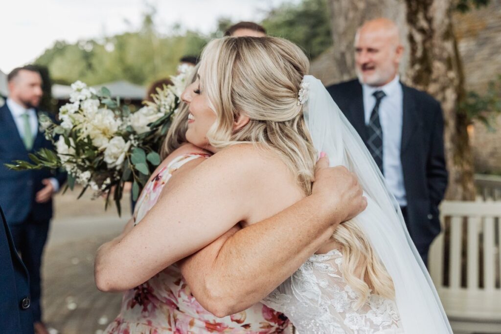 As the bride and her father embrace warmly, a bouquet of white blooms and greenery stands out in the background, with guests in navy suits and floral dresses adding to the joyful at Tithe Barn, Skipton