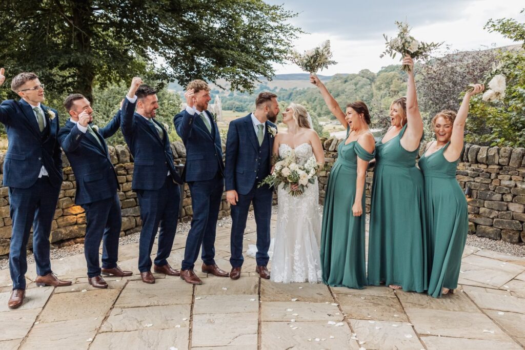 The bridal party celebrates with raised arms, the bride in a flowing white gown and groom in a navy suit at the center, surrounded by bridesmaids in teal dresses and groomsmen at Tithe Barn, Skipton