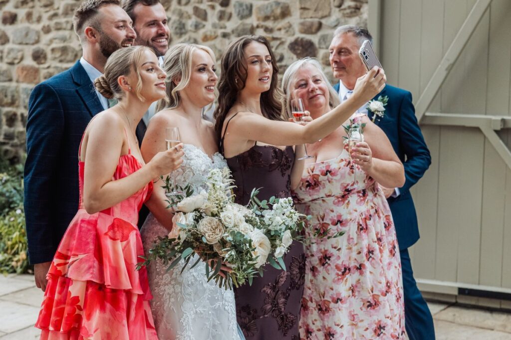 The bride, wearing a lace gown and holding a bouquet of cream blooms, poses with her bridesmaids in vibrant dresses and groomsmen in navy suits, all smiling and capturing the moment at Tithe Barn, Skipton