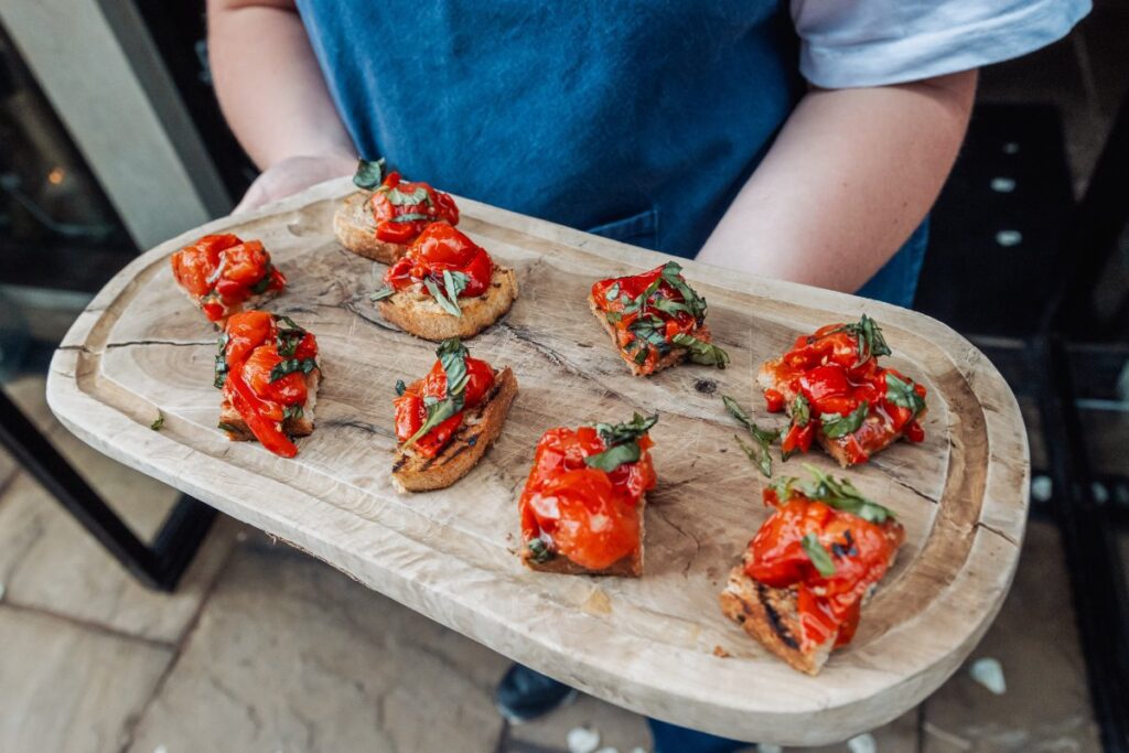 A server presents a wooden platter of vibrant bruschetta appetizers, garnished with fresh basil, at a warmly lit event in a stone-walled courtyard at Tithe Barn, Skipton