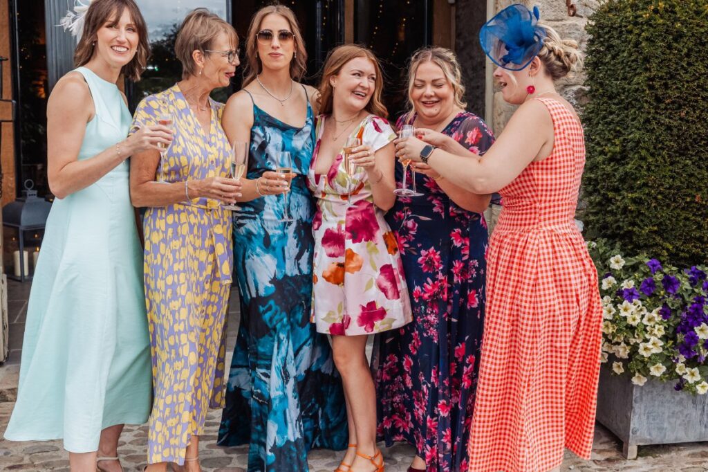 Bridesmaids gather in a sunny courtyard, holding champagne flutes, dressed in vibrant floral and solid-colored gowns, with a stone wall and blooming flowers in the background at Tithe Barn, Skipton