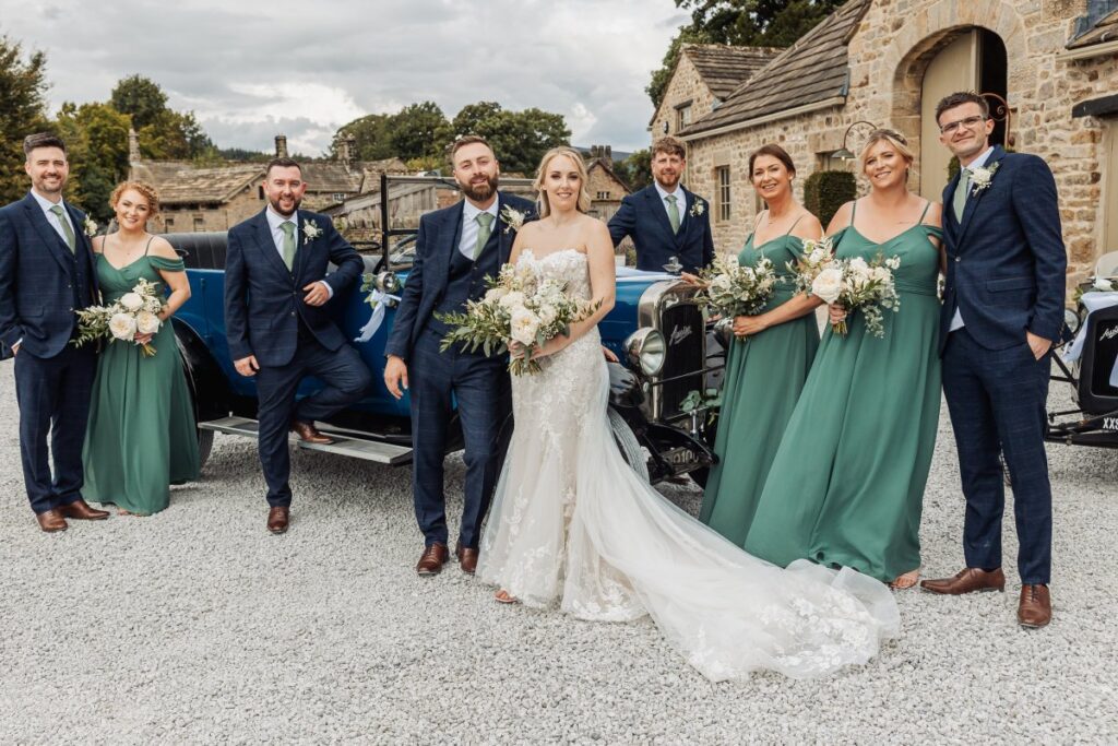 The bridal party poses together in front of a vintage car, with the bride in a flowing white gown and groomsmen in navy suits, while bridesmaids wear elegant teal gowns and hold at Tithe Barn, Skipton
