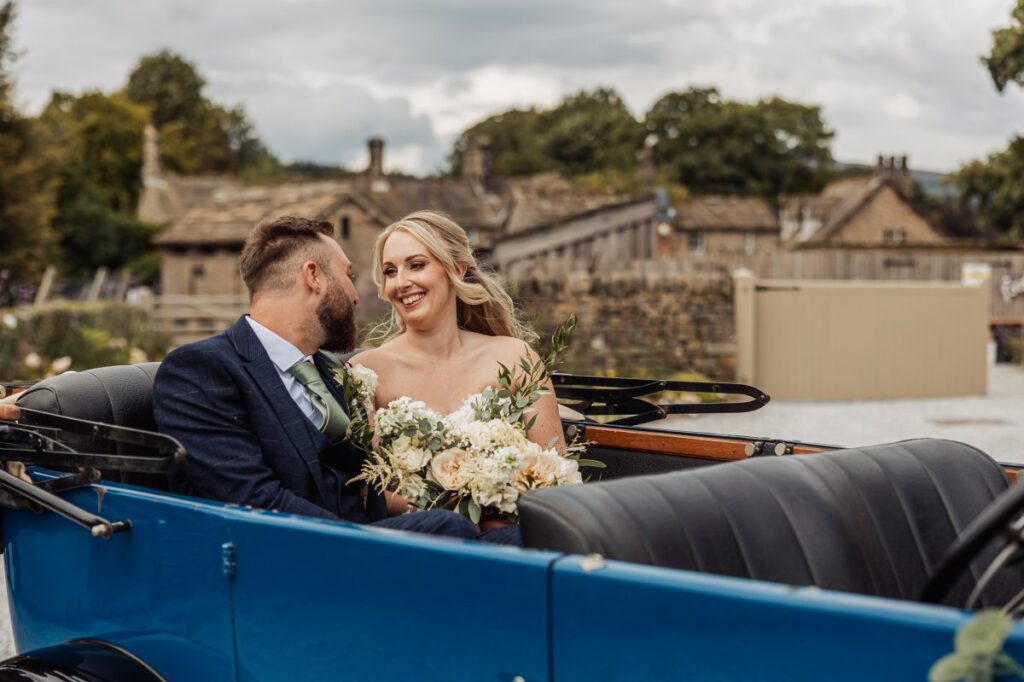 The bride and groom share a joyful moment in a vintage carriage, the bride holding a bouquet of cream and white blooms as they smile at each other against a backdrop of stone buildings at Tithe Barn, Skipton