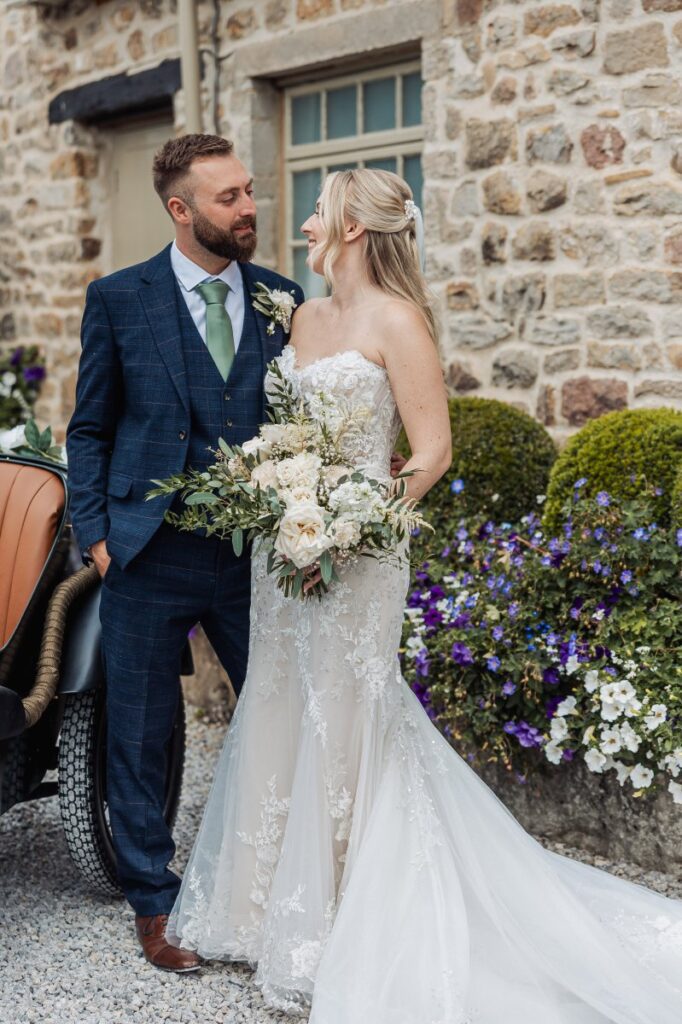 The bride, adorned in a lace-trimmed gown, and groom in a navy suit with a green tie, share a tender moment in front of a stone wall, surrounded by vibrant purple and white blooms at Tithe Barn, Skipton