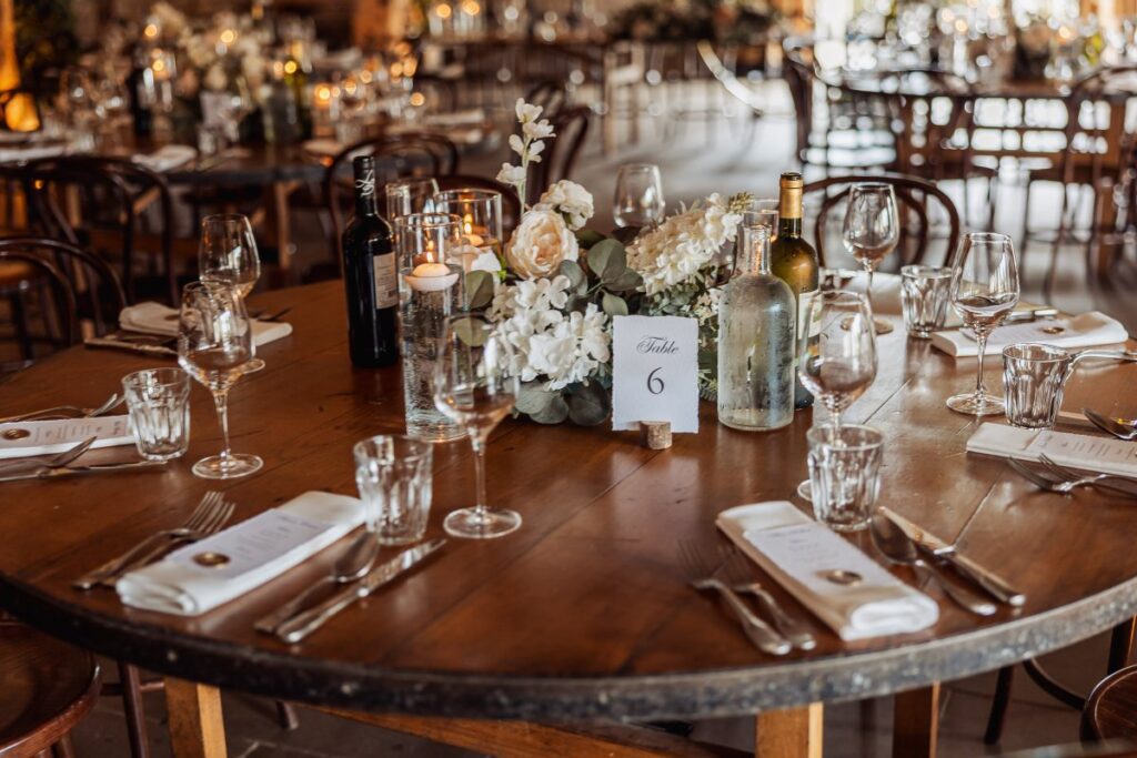 A beautifully set table in a warmly lit wooden hall features a floral centerpiece, candles, and place settings for a wedding reception, with guests seated and ready to dine at Tithe Barn, Skipton