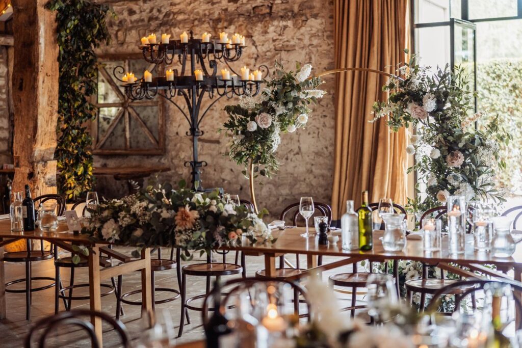 Guests gather under a chandelier, enjoying a candlelit reception with floral arrangements and rustic decor in a warmly lit wooden hall at Tithe Barn, Skipton