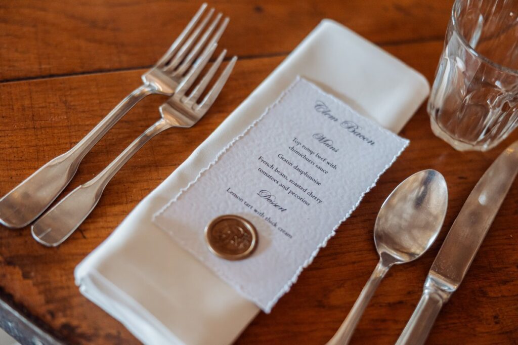 A neatly set table for a wedding reception features a white napkin with a menu card and a copper coin, flanked by silverware and a clear glass, all arranged on a warm wooden surface at Tithe Barn, Skipton
