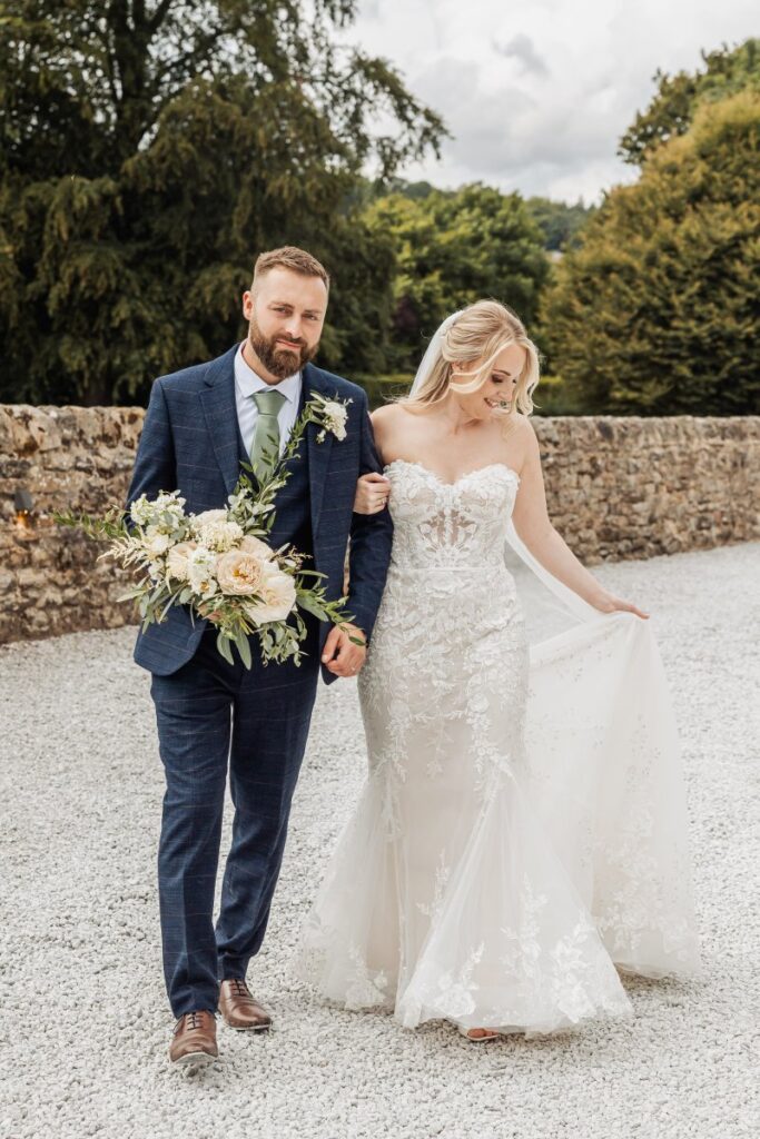 The bride and groom walk hand-in-hand through a walled garden, the bride in a flowing lace gown and the groom in a navy suit, with a bouquet of cream and green blooms adding a touch at Tithe Barn, Skipton