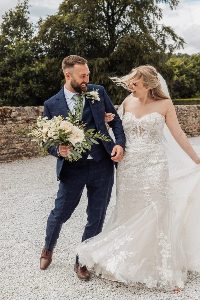Bride and groom walk across the gravel at Tithe Barn Bolton Abbey, the groom carrying her bouquet as her veil catches the wind.