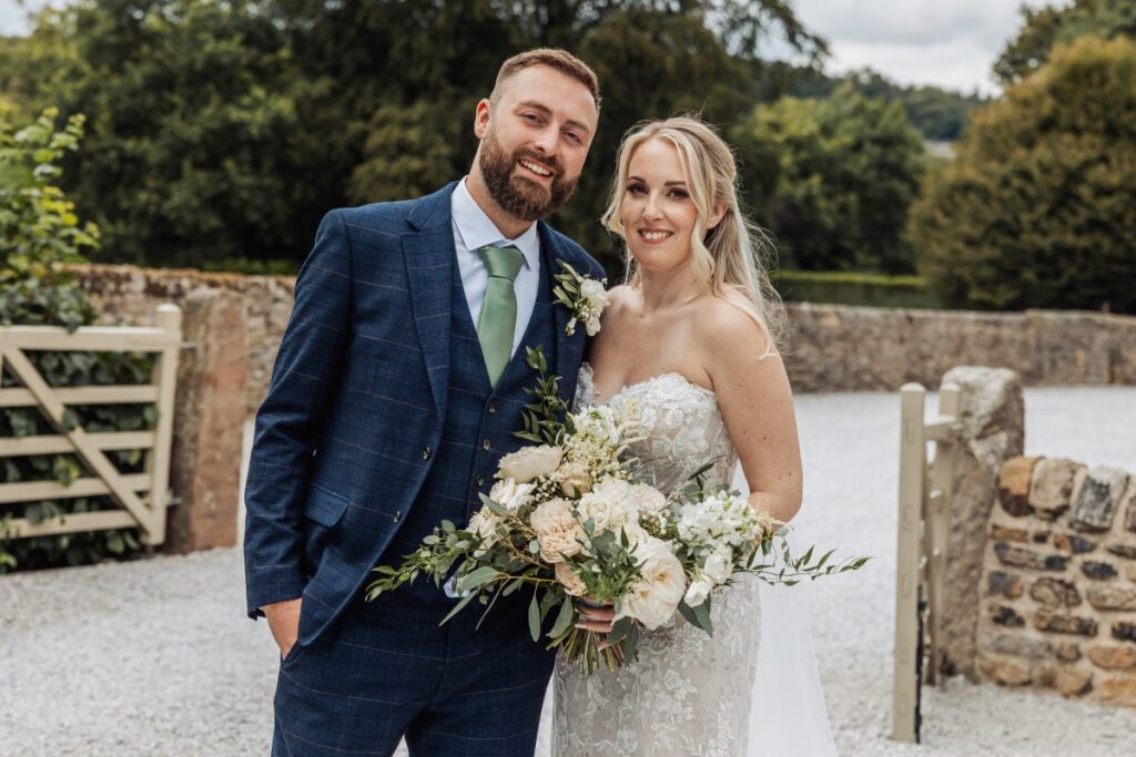 Portrait of the bride and groom on the gravel outside Tithe Barn Bolton Abbey, her bouquet of cream roses and eucalyptus held between them.