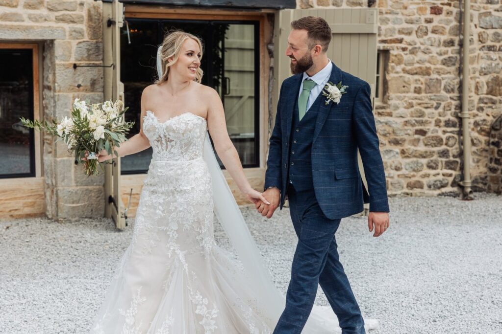 As they walk hand-in-hand, the bride in a lace gown and the groom in a navy suit with a boutonniere share a joyful moment in front of a rustic stone building with a vase of cream at Tithe Barn, Skipton