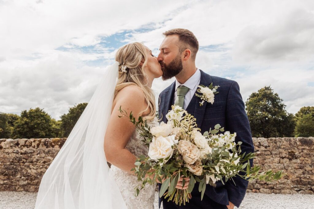 The bride and groom share a tender kiss, their faces close as the bride holds a lush bouquet of cream and greenery, while the groom wears a navy suit with a floral boutonniere, set at Tithe Barn, Skipton