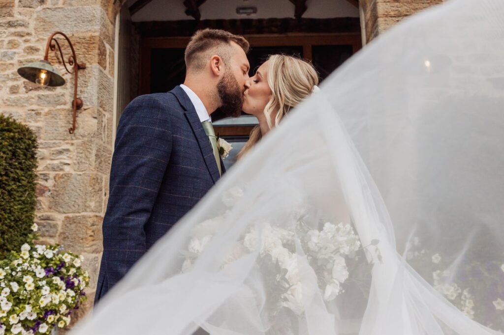 A bride in a flowing white gown and veil shares a tender kiss with her groom in a navy suit, framed by a stone archway and a wall of vibrant flowers at Tithe Barn, Skipton