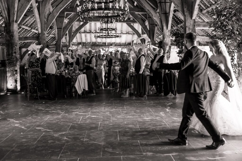 The couple enter the reception at Tithe Barn Bolton Abbey through a tunnel of guests waving napkins, in black and white.