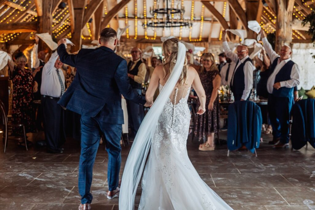 Bride and groom walk into the reception at Tithe Barn Bolton Abbey as guests wave white napkins overhead.
