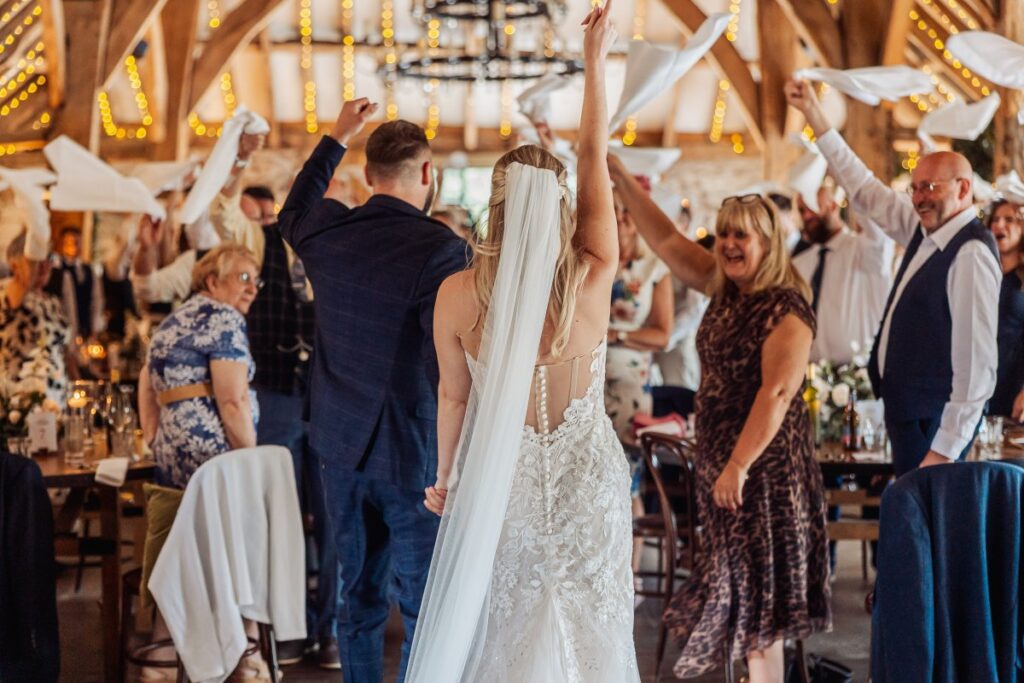 The bride raises her arms as she walks into the reception at Tithe Barn Bolton Abbey, guests waving napkins on both sides.