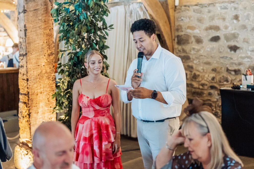 During speeches, a man in a white shirt holds a microphone, while a woman in a vibrant red dress stands beside him, as guests in a warmly lit stone-walled hall look on at Tithe Barn, Skipton